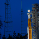 A orange and white rocket stands up next to its scaffolding highlighted by surrounding floodlights. It's nosecone sits directly in front of a full moon.